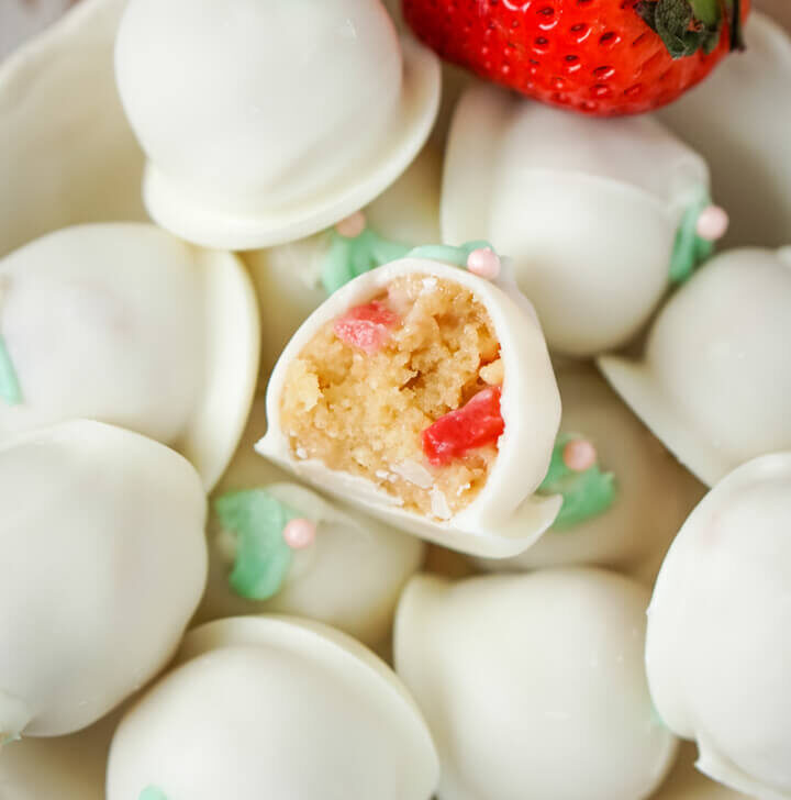 A bowl full of strawberry cheesecake truffles with 2 fresh strawberries to the side.