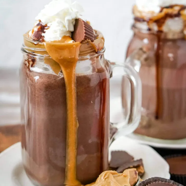 Two mugs of peanut butter hot chocolate garnished with whipped cream, peanut butter topping, chocolate syrup and peanut butter cup candies.