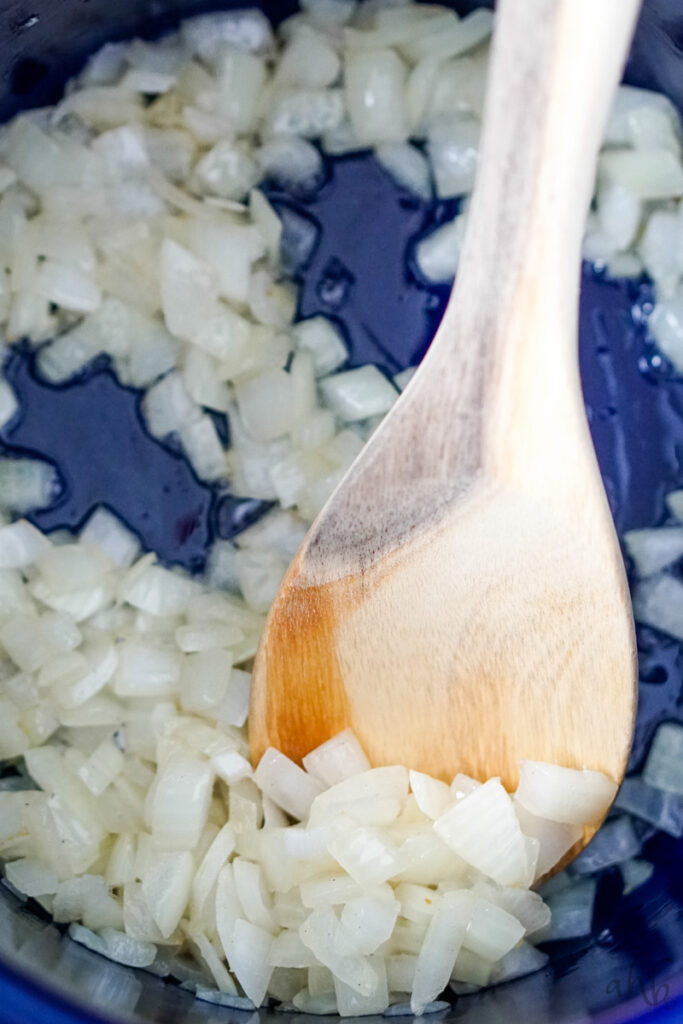 Diced onion sautéing in bacon grease being stirred by a wooden spoon in a blue stockpot.