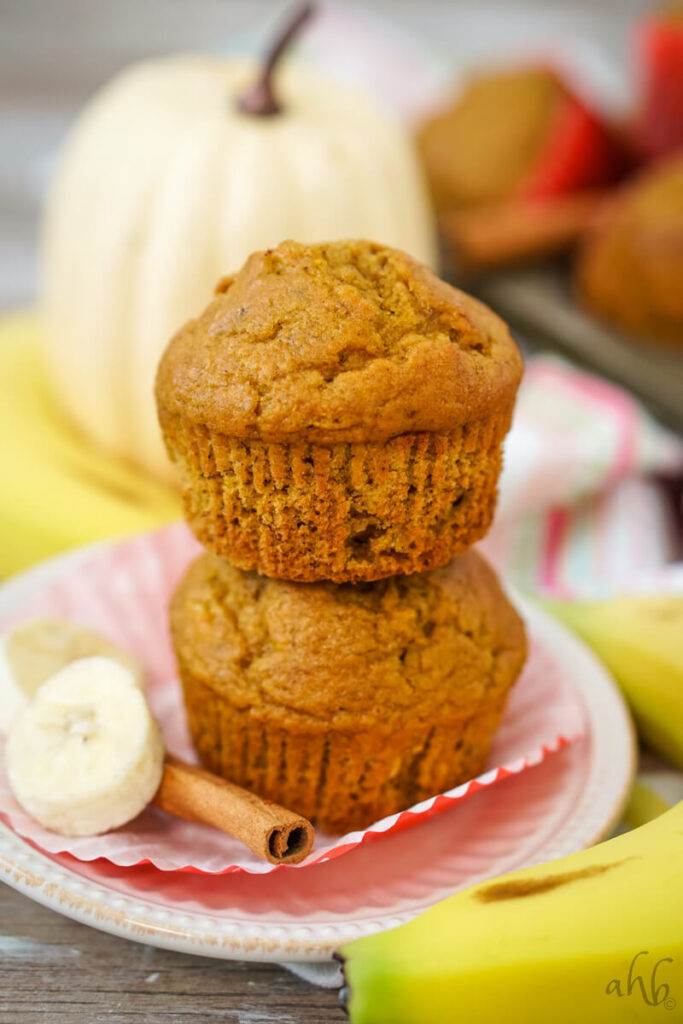 An unwrapped Pumpkin Banana Muffins on top of another unwrapped muffin next to sliced banana and a cinnamon stick. 