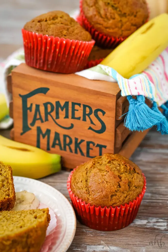 In the foreground, a pumpkin banana muffin in a red paper muffin liner next to a sliced open muffin and a banana. In the background two pumpkin banana muffins in red liners next to a banana on a wooden box that says Farmers Market.