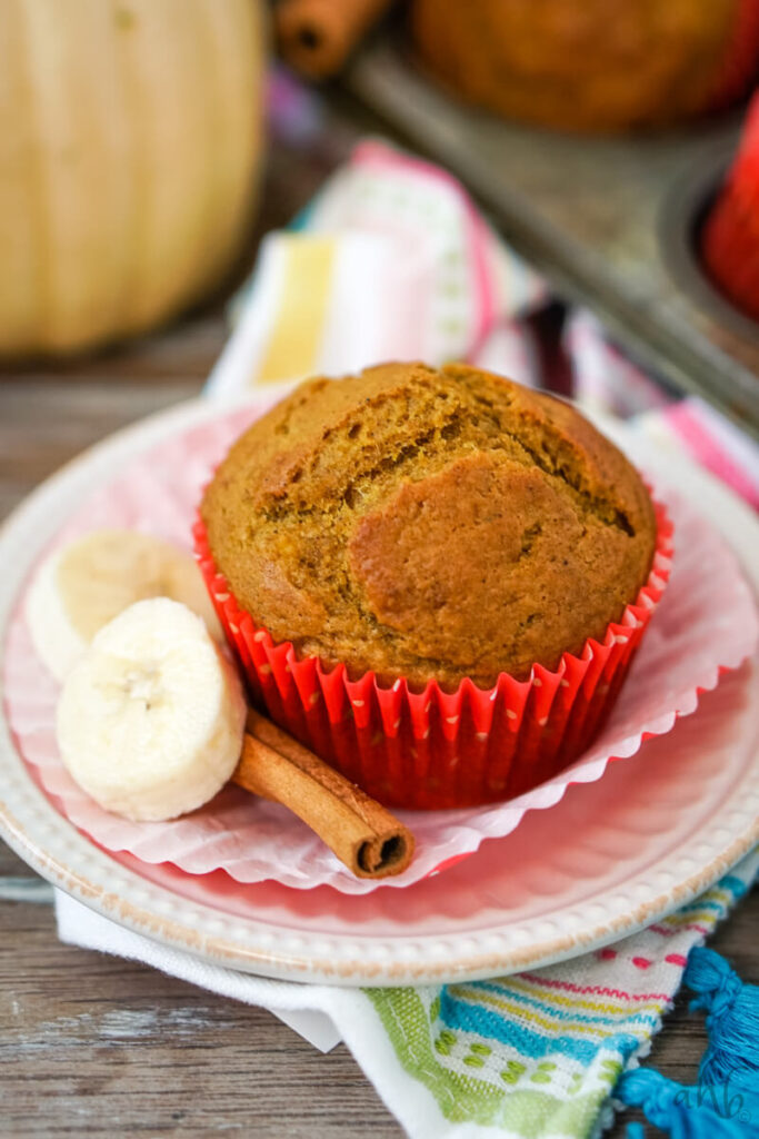 A Pumpkin banana muffin in two paper muffin liners with two slices of bananas and a cinnamon stick on a decorative white plate. 