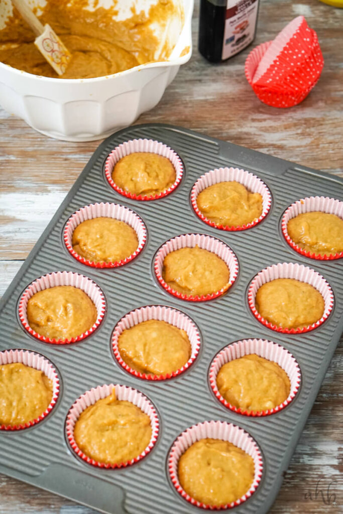 The batter from the white mixing bowl is distributed into twelve muffin tins lined with paper muffin liners. 