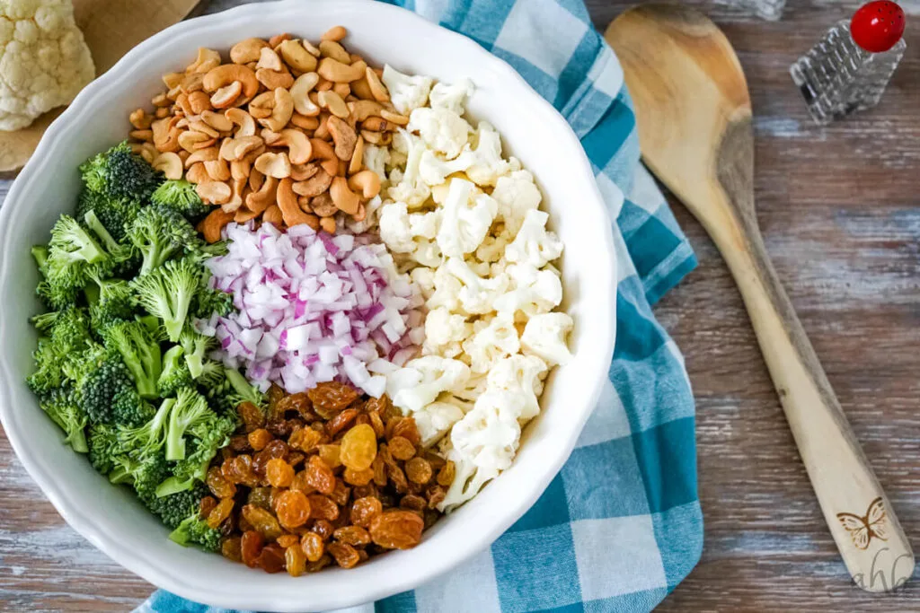 A large white bowl full of chopped broccoli, cashews, cauliflower, golden raisins, and red onions.