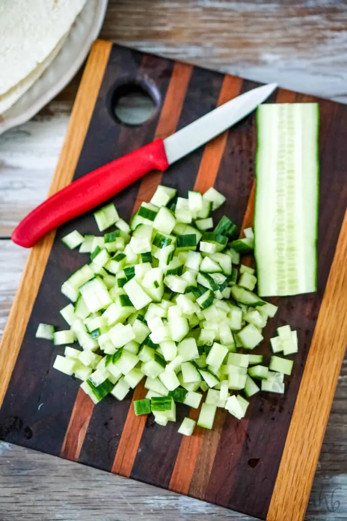 Half of an English cucumber, diced, next to an intact cucumber half on a wooden cutting board with a red handled knife.