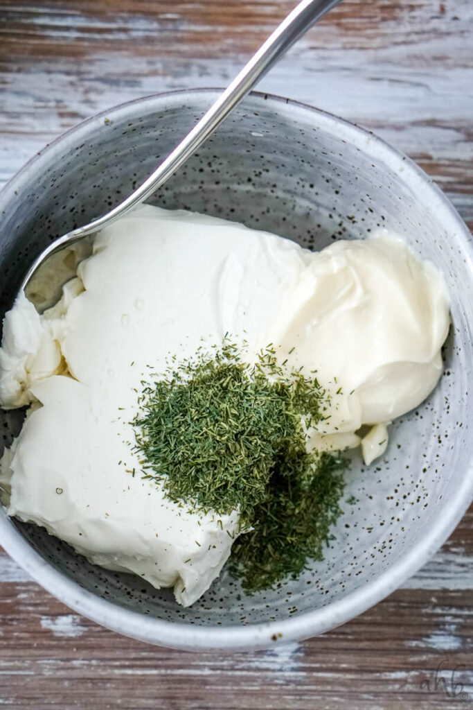 Cream cheese, mayo, and dill weed is mixed together with a spoon in a gray mixing bowl.