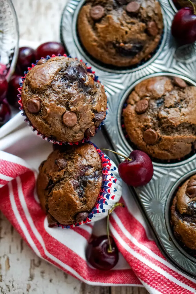 Two Cherry Chocolate muffins next to a pan of muffins and cherries.
