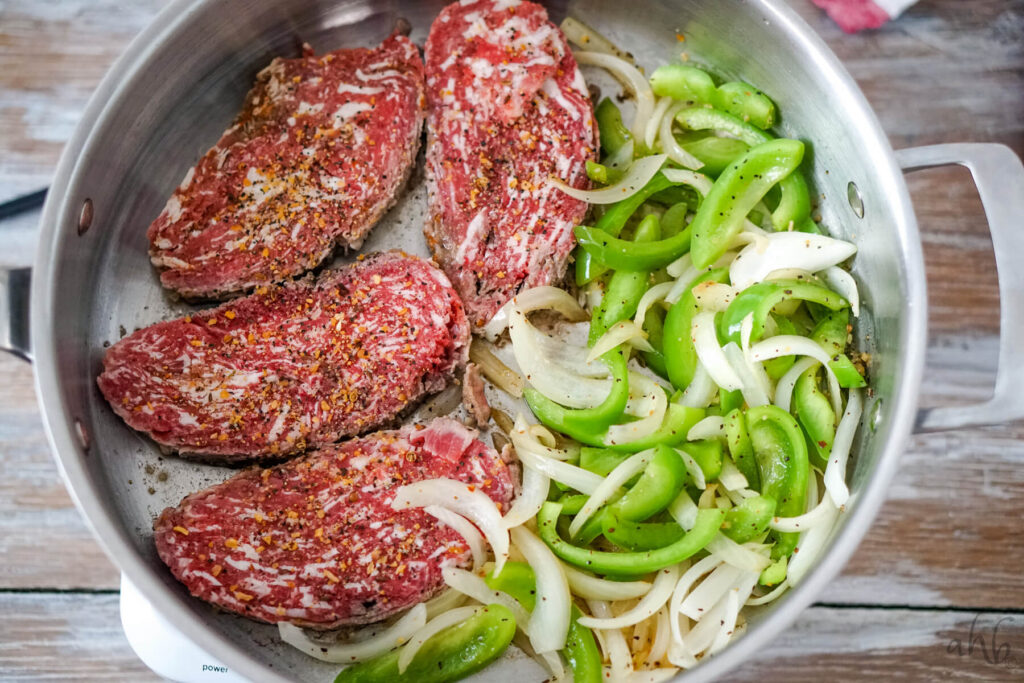 Four steaks cook in a sauté pan with slices of green pepper and onion.