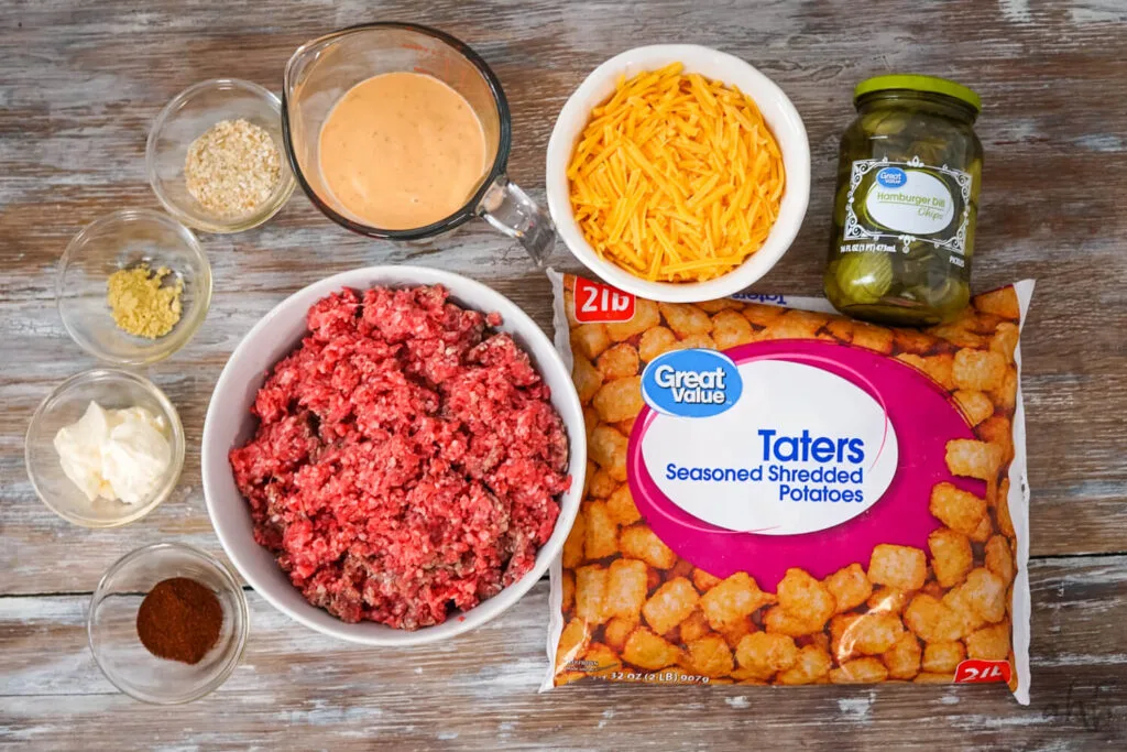 A 32 oz. bag of frozen tater tots, a jar of hamburger dill pickle slices, shredded cheddar cheese, thousand island dressing, 2 lbs. of raw ground beef, paprika, mayo, dried ground mustard, and dried minced onion on a wooden table.