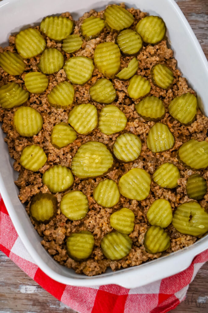 Browned ground beef is evenly spread in the bottom of a white baking dish. Pickle slices are evenly placed on top.