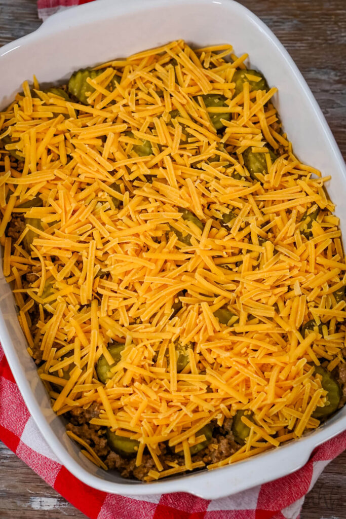 Ground beef and pickles are topped with shredded cheddar cheese in a white baking dish.