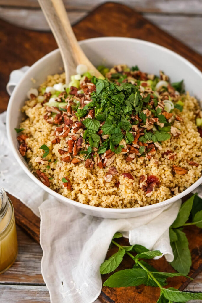 A white bowl of cranberries and couscous topped with toasted pecans, green onions, and mint leaves being stirred with a wooden spoon.