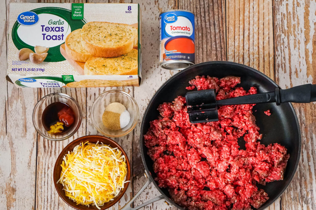 A box of Garlic Texas Toast, Condensed Tomato soup, Worcestershire sauce, yellow mustard, and ketchup, onion powder, salt, and garlic powder, shredded colby jack cheese, and broken up raw ground beef sitting in a medium black skillet.