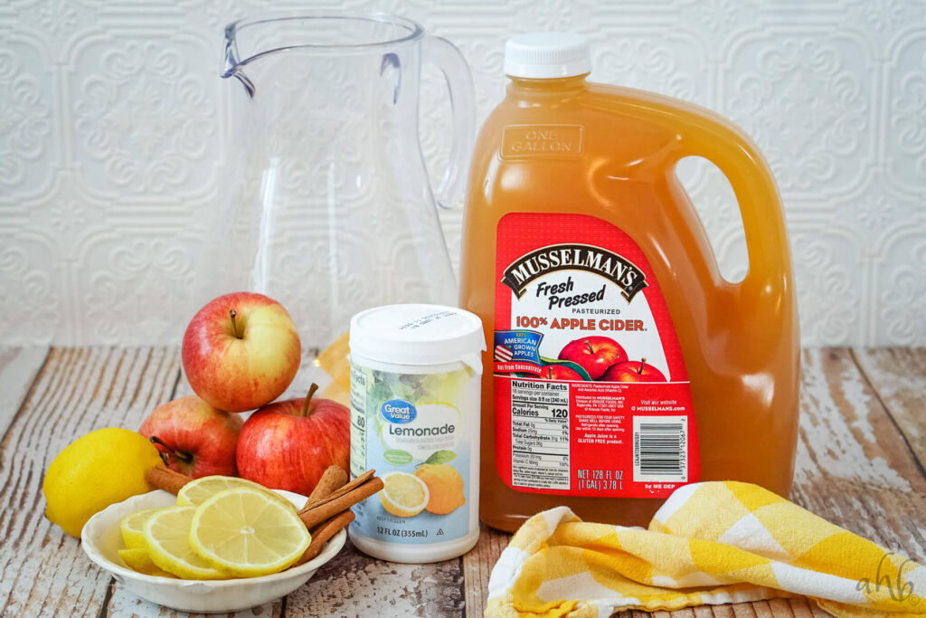 A large, glass pitcher, three red apples, a lemon, lemon slices, cinnamon sticks, Lemonade concentrate, and a gallon of apple cider.