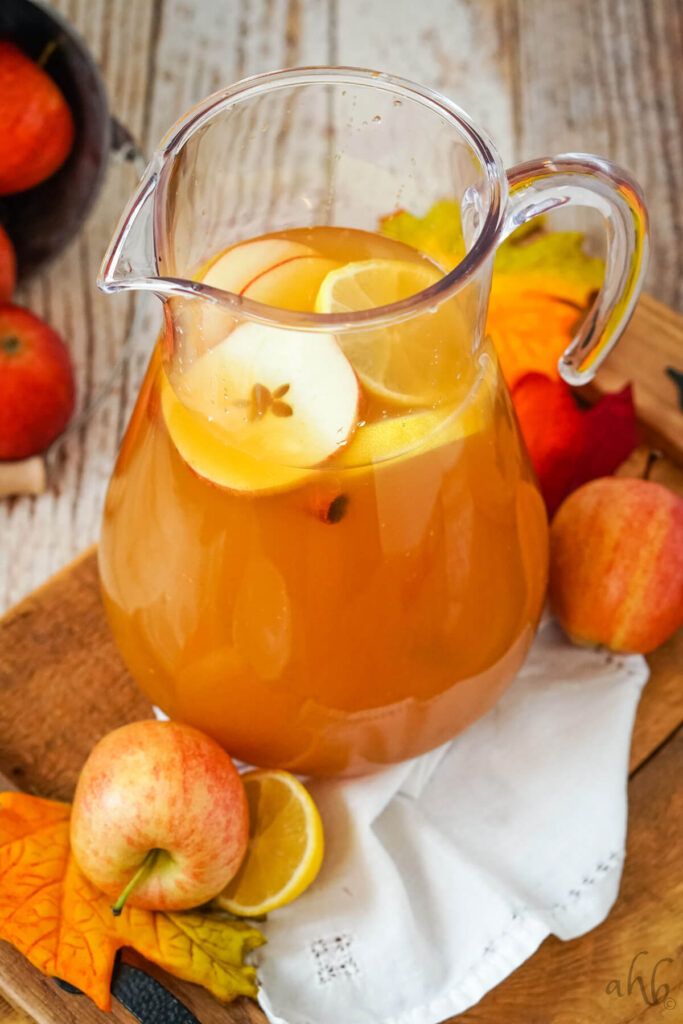 A glass pitcher full of Lemonade Cider garnished with sliced apples, lemons, and cinnamon sticks surrounded by apples.