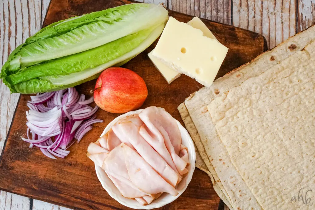 A head of romaine lettuce, a red apple, two blocks of swiss cheese, thinly sliced red onion, sliced turkey, and four large tortillas on a wooden cutting board.
