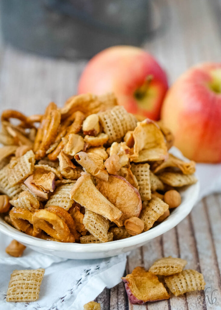 A serving of Carmel Apple Chex Mix in a white dish in front of two apples.