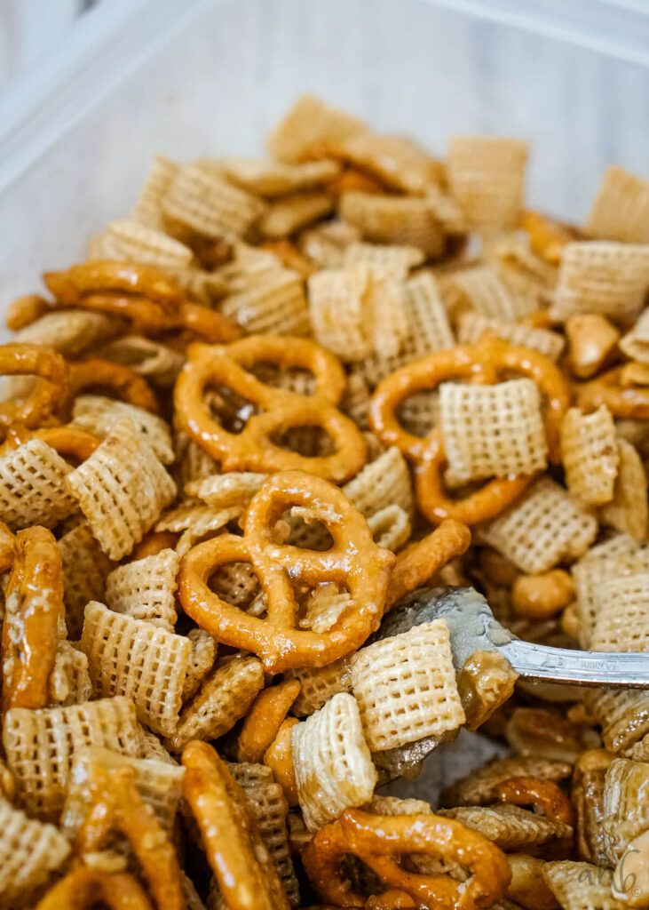 A mix of Chex cereal, pretzels, and peanuts covered in the butter and brown sugar mixture stirred by a spoon.