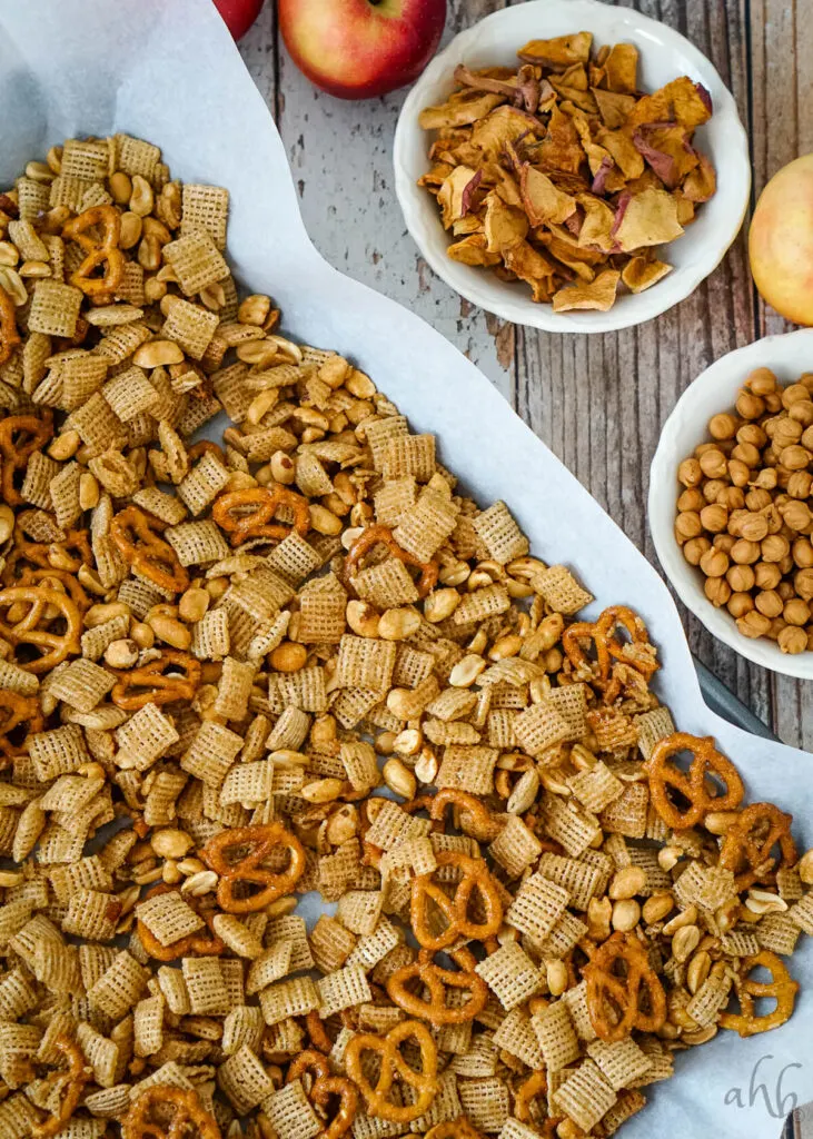 The cereal mixture is laid out on a baking tray to cool next toa bowl of apple chips and a bowl of caramel chips.