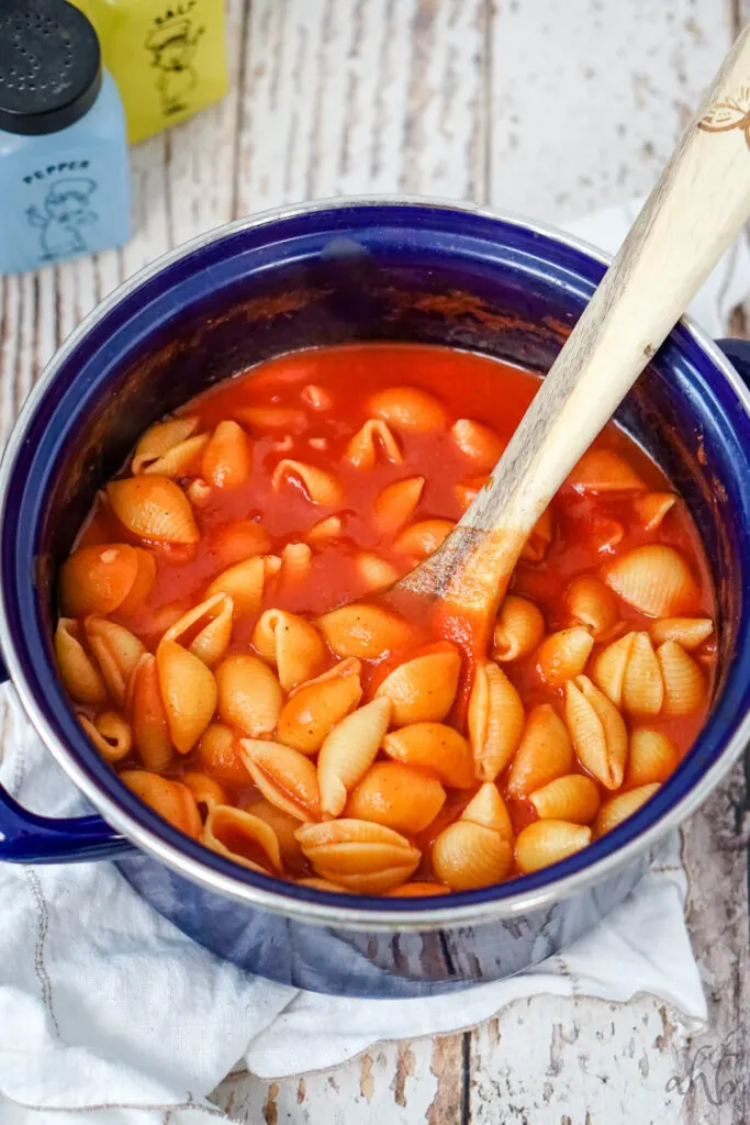 The seasoned Old Fashioned Tomato Macaroni is stirred with a wooden spoon in a blue stockpot.
