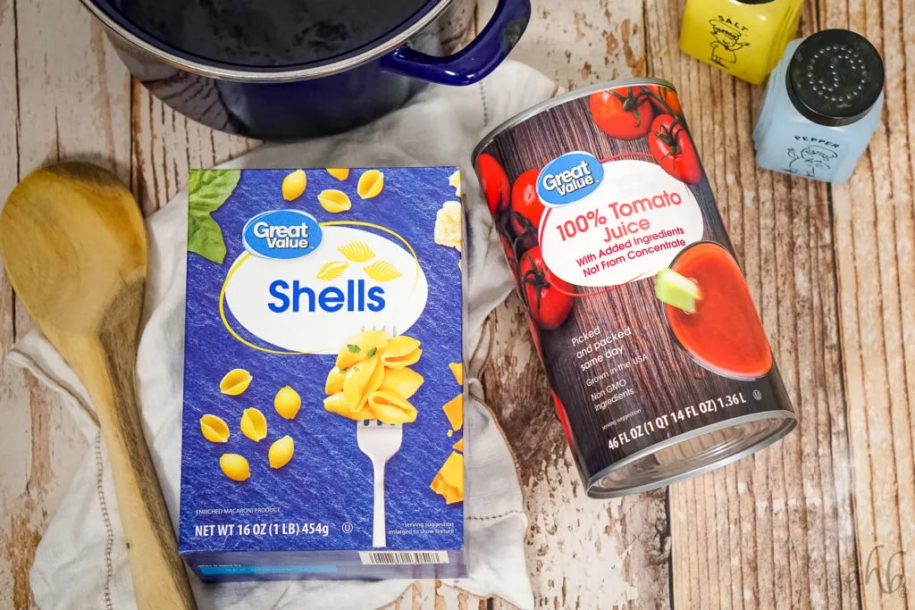 A box of shell pasta, a can of tomato juice, slat, pepper, a wooden spoon, and a blue stockpot.