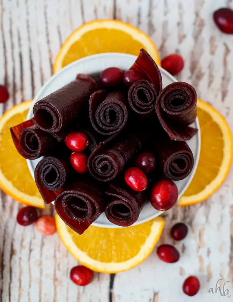A white bowl of cranberry fruit leather rolls with orange slices and cranberries. 