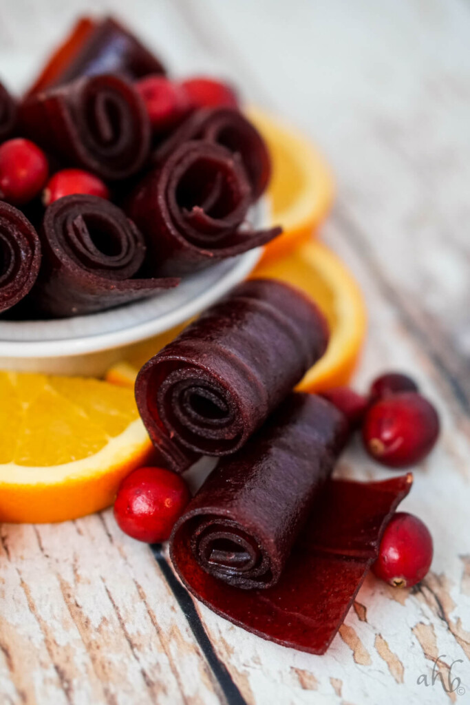Two Cranberry orange fruit leather rolls with several cranberries and orange slices next to a bowl of cranberry orange fruit leather rolls. 