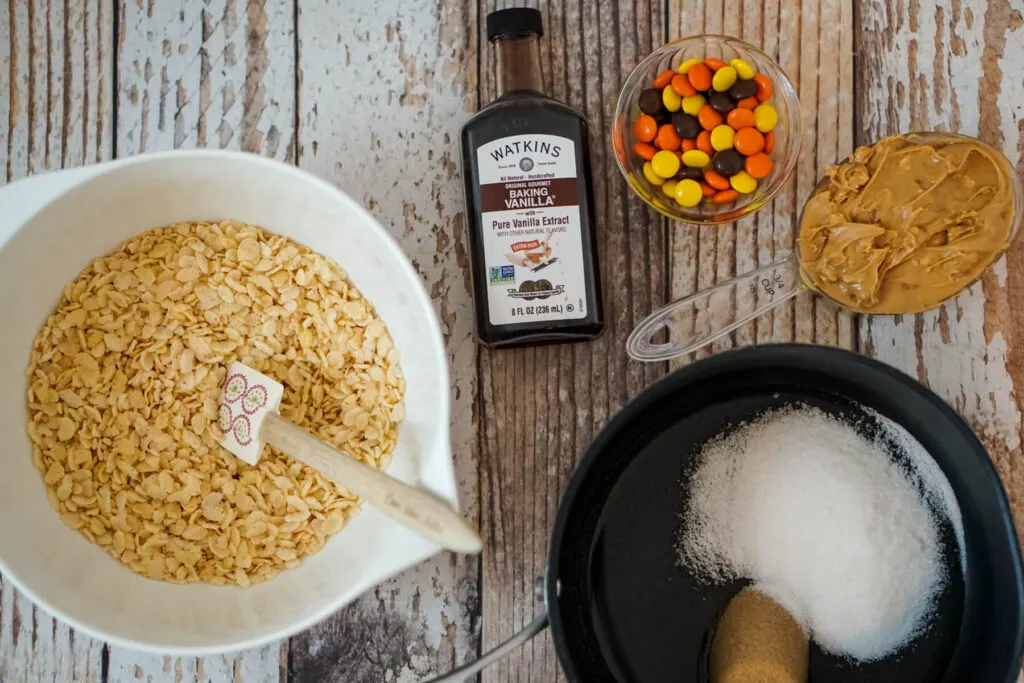 A bowl of Rice Krispies, a bottle of vanilla extract, a serving of Reese's pieces, 3/4 cup of peanut butter, and a small saucepan of corn syrup, brown sugar, and granulated sugar.