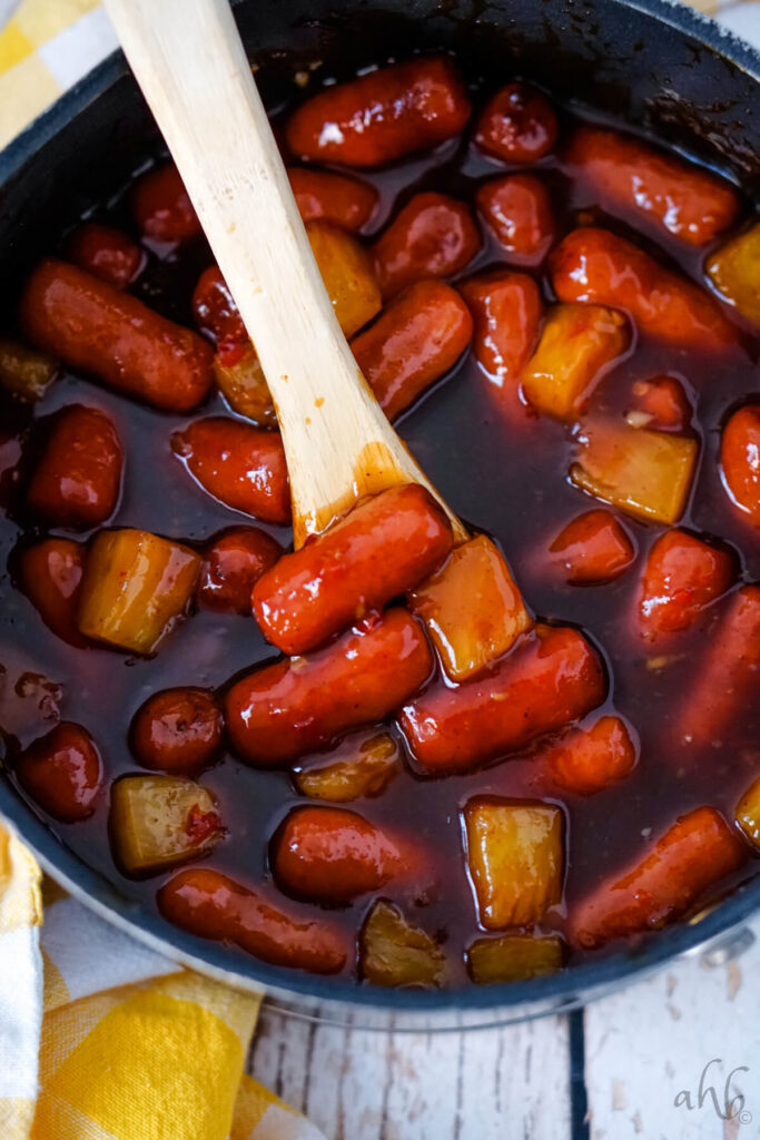 A pot of Hawaiian Lit'l Smokies stirred with a wooden spoon. 