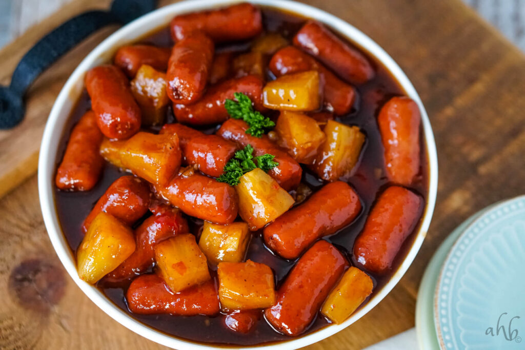 A bowl of little smokies with pineapple and sauce on a serving tray. 