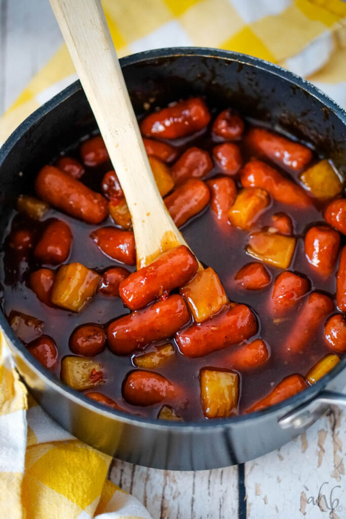 A pot of Hawaiian Lit'l Smokies stirred with a wooden spoon.