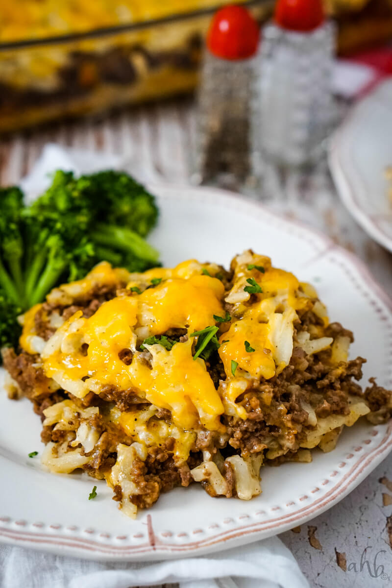 A serving of Hamburger Hashbrown Casserole is served with cooked broccoli on a white plate.