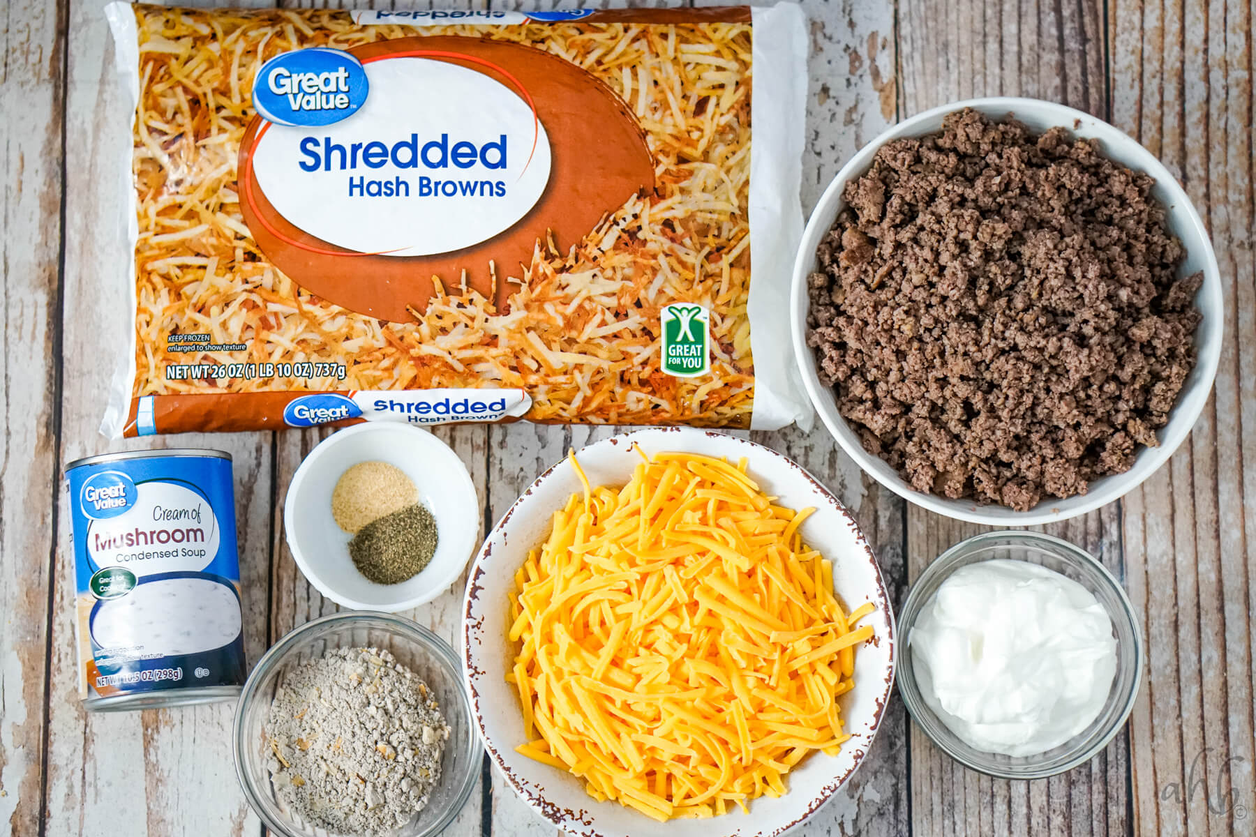 A bag of shredded hashbrowns, a bowl of browned ground beef, a bowl of shredded cheddar cheese, pepper, garlic powder, a can of cream of mushroom soup, dry onion soup mix, and sour cream.