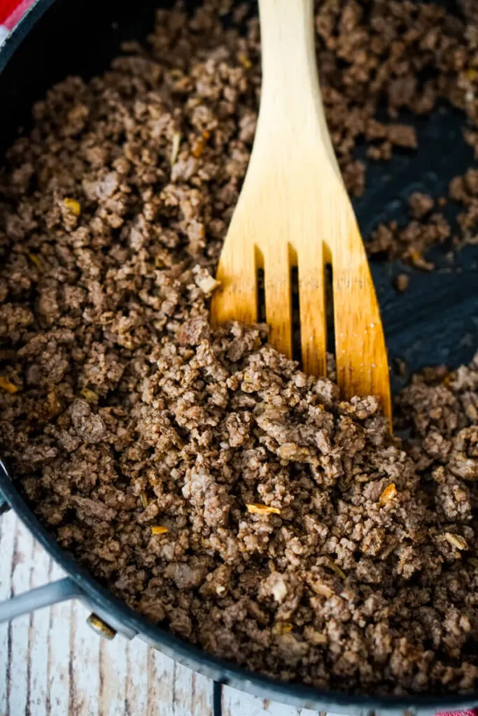 Browned ground beef is stirred by a wooden spoon in a skillet.