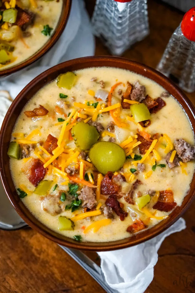 A bowl of Slow Cooker Bacon Cheeseburger Soup served with shredded cheddar cheese, sliced pickles, and bacon pieces.