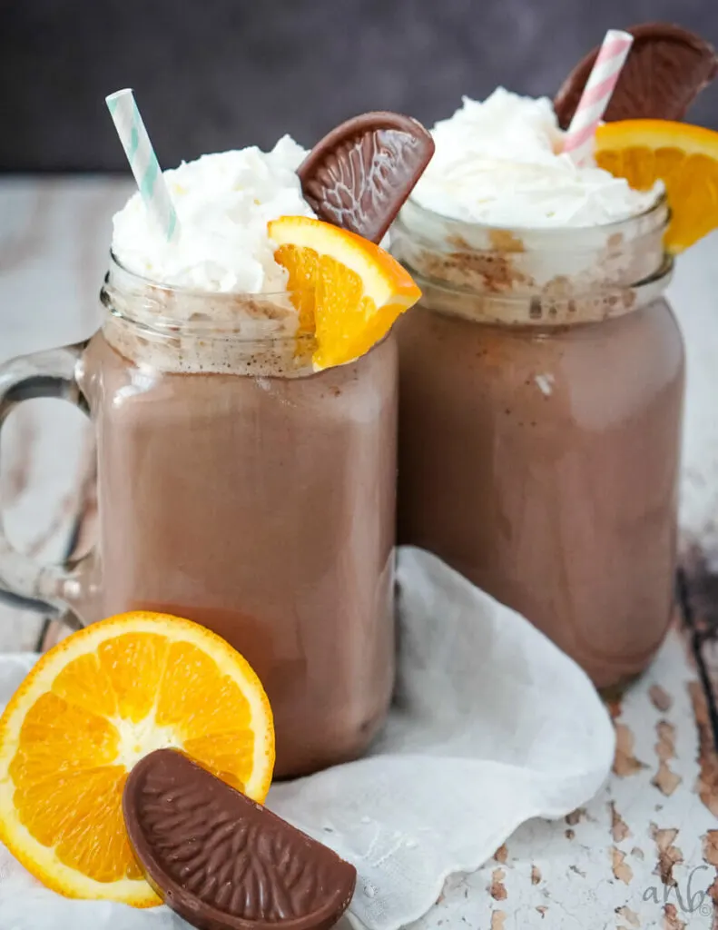 Clear glass mugs full of orange hot chocolate with striped straws in them. An orange and a chocolate in front of the mugs. 