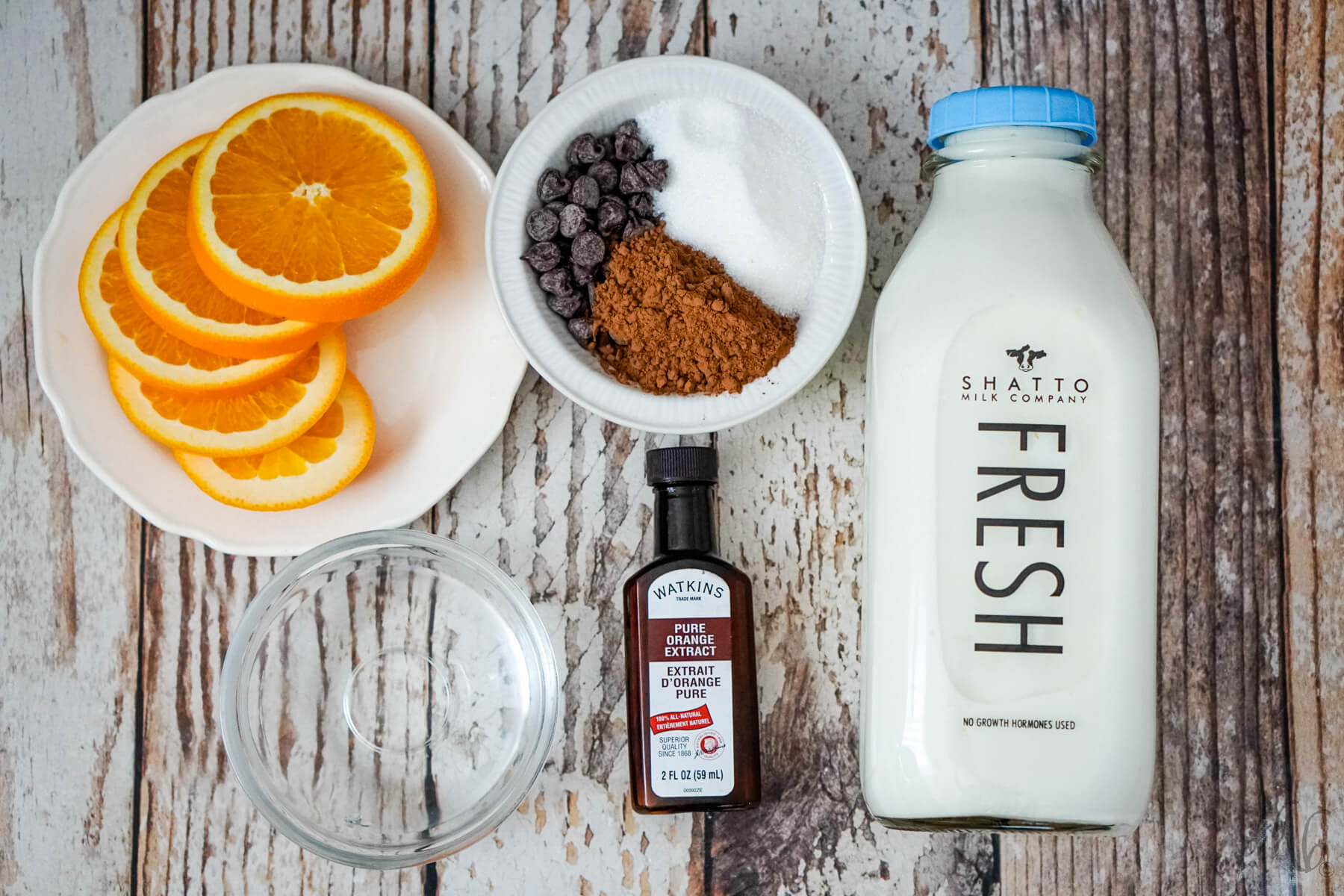 Slices of orange on a white plate, a bowls of granulated sugar, cocoa powder, and semi-sweet chocolate chips in a white bowl, a bowl of water, a bottle of orange extract, and a bottle of Shatto milk.