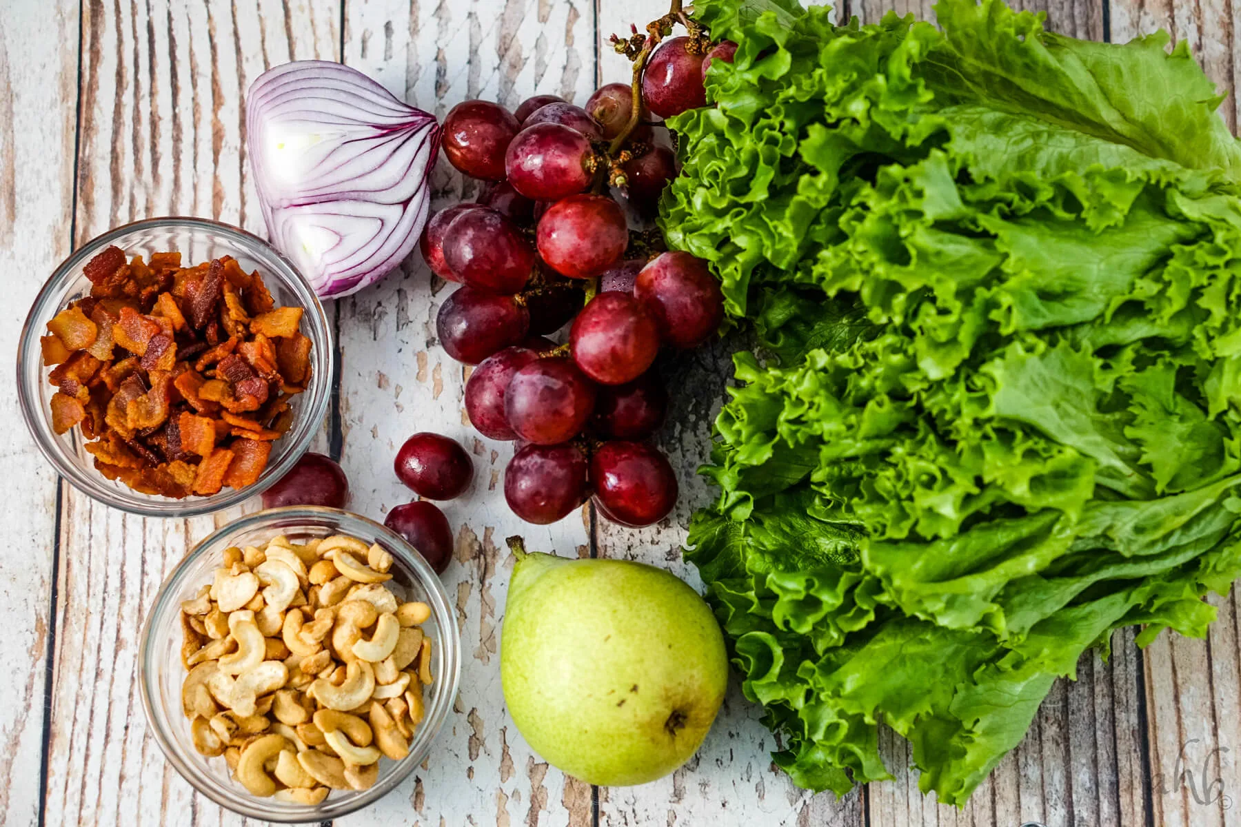 A head of lettuce, a pear, some red seedless grapes, half of a red onion, a bowl of bacon pieces and a bowl of cashew halves and pieces sit next to each other.