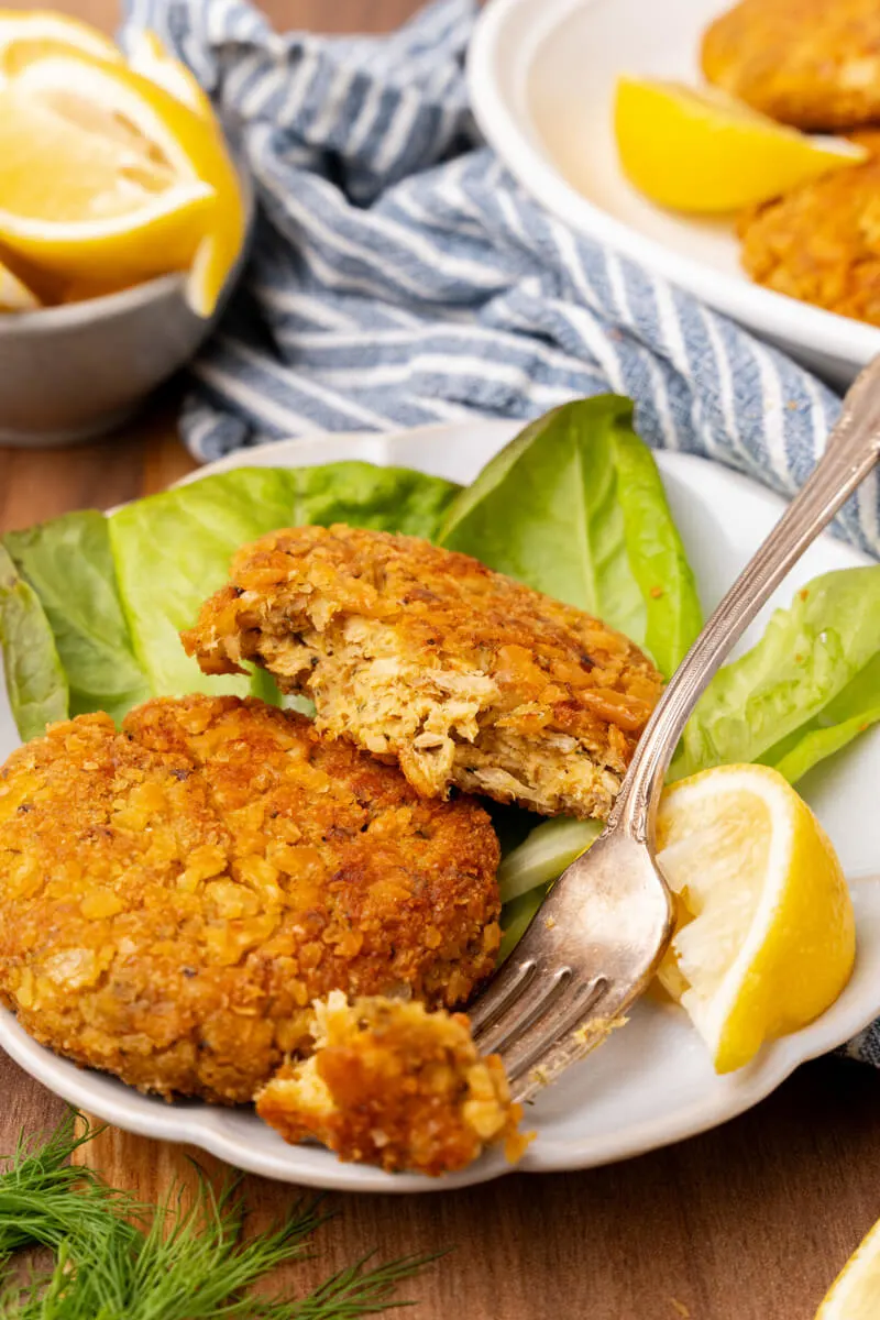 A plate of two salmon patties served with a lettuce leaf and a lemon slice. One of the patties is half eaten, and a fork takes a bite sized piece from the plate.