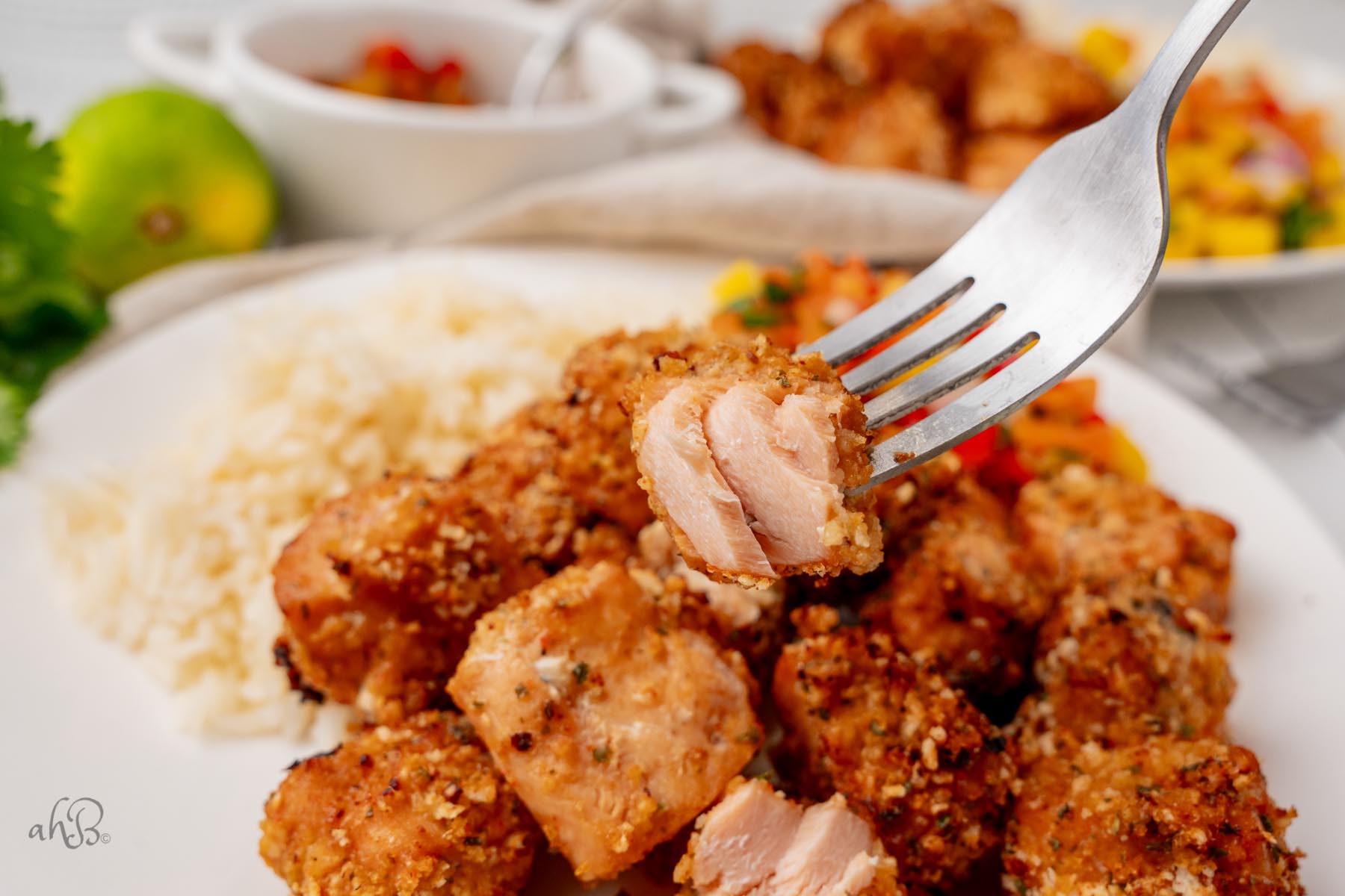 Half of a salmon bite is held on a fork over a plate of salmon bites and rice.